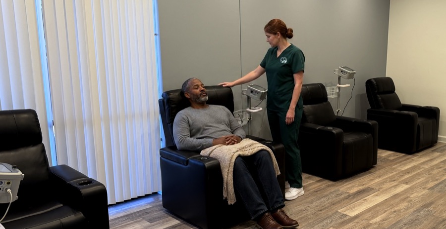 Our treatment room: a nurse in Brain Recovery Centers scrubs beside a visitor resting in a recliner, with a blood-pressure monitor on a stand beside each chair.