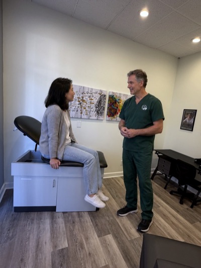 A clinician in Brain Recovery Centers scrubs talks with a visitor seated on the exam table.