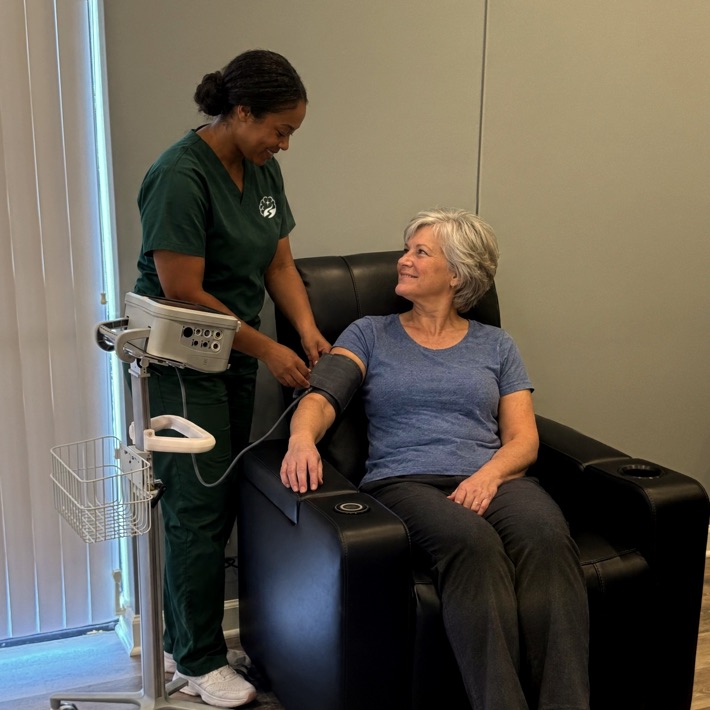 A nurse in Brain Recovery Centers scrubs checks the blood pressure of a visitor seated in a treatment recliner.